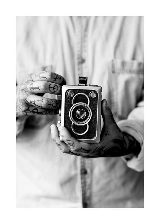  – Sort-hvidt fotografi af et vintagekamera, der holdes af en mand med tatoverede hænder