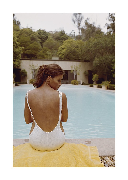  – Fotografi af en kvinde, der sidder på et håndklæde foran en swimmingpool, iført en hvid badedragt