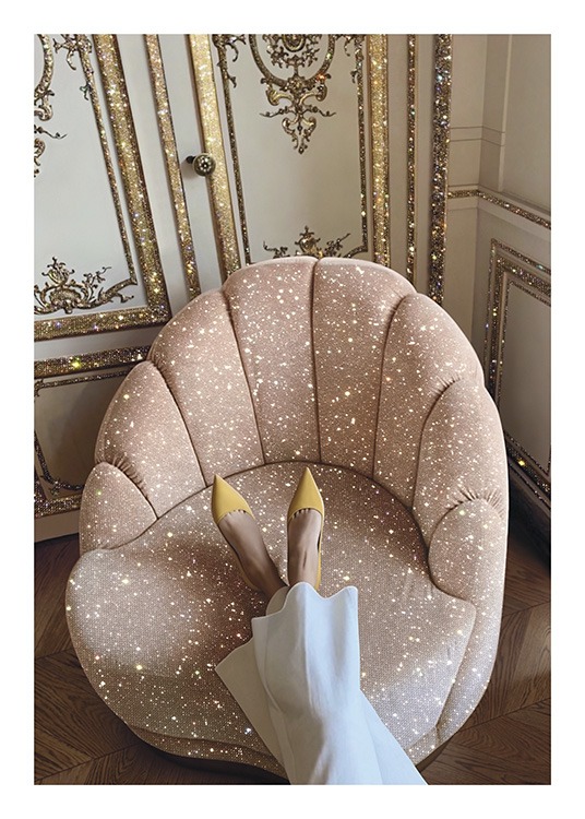  – Fotografi af en lyserød stol med glimmer, som en kvinde har lagt fødderne på, foran en gylden og hvid væg
