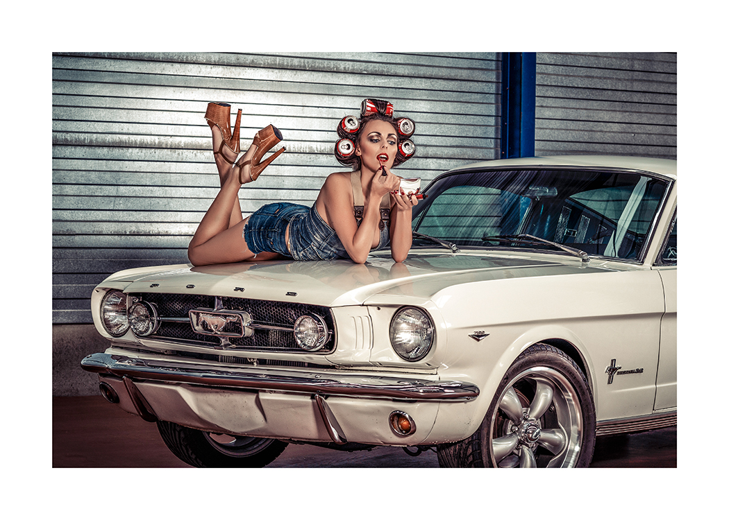  – Fotografi af en kvinde med sodavandsdåser i håret, der ligger på kølerhjelmen af en vintagebil og tager læbestift på