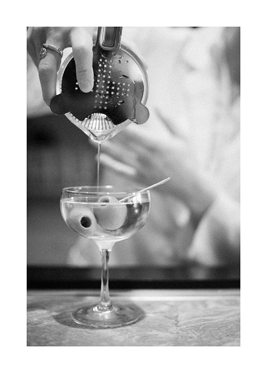  – Sort-hvid fotografi af en martini i en drinkshaker, der hældes ned i et glas med oliven