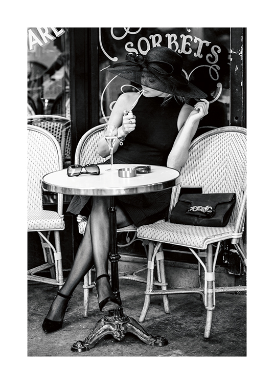  – Sort-hvidt fotografi af en kvinde i en stor hat, der sidder uden for en café og tænder en cigaret